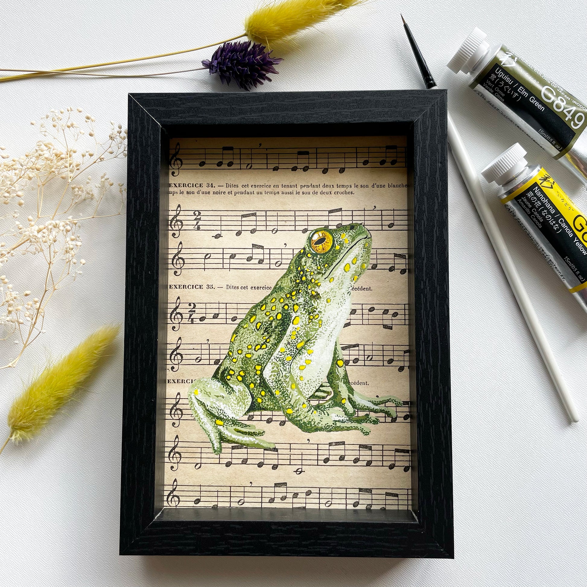 Grenouille verte  Peinture originale réalisée à la gouache extra-fine sur une page de partition de musique ancienne.  Pièce unique  Le cadre mesure :  11,8 cm de largeur sur 16,8 cm de hauteur (mesures extérieures) profondeur 2,5 cm Fabriqué en France 