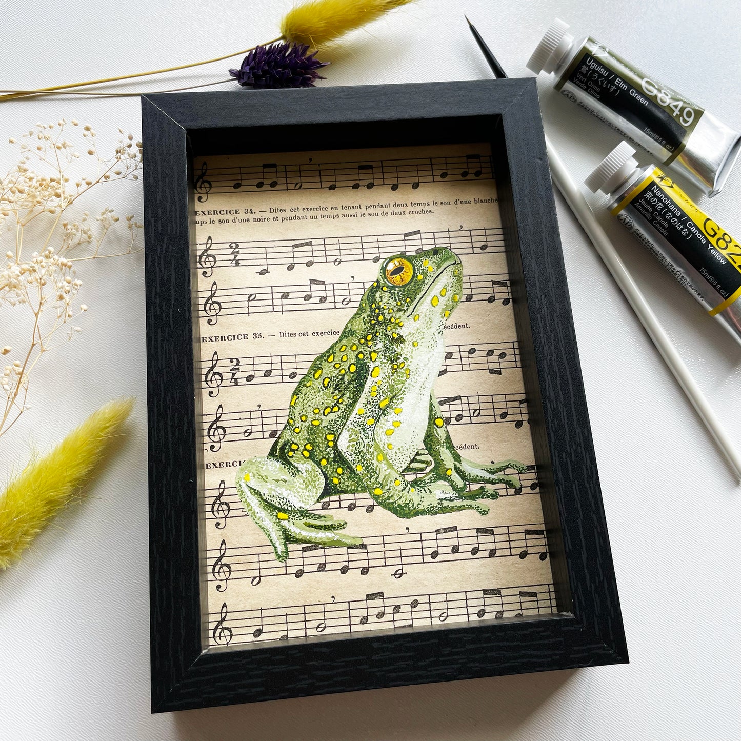 Grenouille verte  Peinture originale réalisée à la gouache extra-fine sur une page de partition de musique ancienne.  Pièce unique  Le cadre mesure :  11,8 cm de largeur sur 16,8 cm de hauteur (mesures extérieures) profondeur 2,5 cm Fabriqué en France 